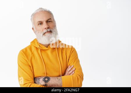 Cool and stylish senior man with tattoos and beard, cross arms on chest, looking confident and unbothered, wearing orange hoodie, standing over white Stock Photo