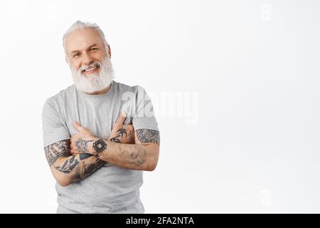 Stylish mature man with tattoos and long beard cross arms on chest, smiling and looking candid at camera. Authentic carefree senior man standing Stock Photo