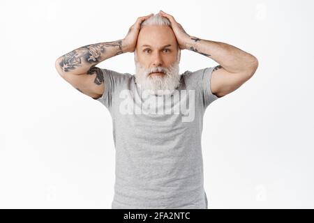 Troubled and worried old man with tattoos, holding hands on head and look confused, panicking or feeling nervous about accident, standing alarmed and Stock Photo