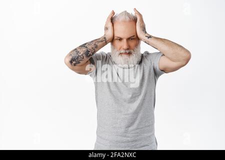 Annoyed and distressed old man with tattoos, holding hands on head with troubled upset face, looking down bothered, fed up, tired of problems Stock Photo