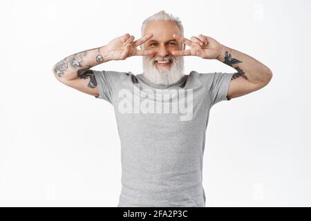 Funny and happy senior guy with tattoos showing disco peace v-signs near eyes, smiling and looking positive at camera, standing in gray t-shirt Stock Photo