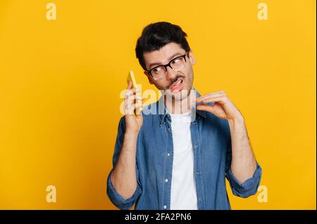 Phone call by cellphone. Attractive charismatic stylish guy has an unpleasant phone conversation, ignores the interlocutor, makes blah blah gesture, stands on isolated orange background, looks away Stock Photo