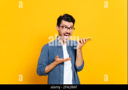 Emotional caucasian stylish guy with glasses, holding smartphone in hand, screaming during phone call or recording audio message, stand on an isolated orange background. Audio messaging Stock Photo