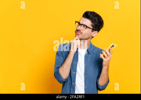Confused puzzled caucasian thoughtful guy, stylishly dressed and with glasses, holding a smartphone in his hand, looking pensively to the side, standing an isolated orange background Stock Photo