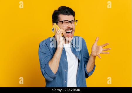 Unpleasant phone conversation. Irritated angry caucasian young man yelling during phone conversation, gesturing with hand, stands on an isolated orange background Stock Photo