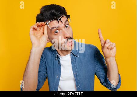 Amazed surprised caucasian guy in an orange t-shirt, shocked looking at the camera with his glasses raised, shows IDEA gesture, standing on an isolated orange background Stock Photo