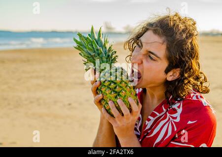 Young Long Hair Man in Red Hawaiian Shirt Opening Mouth to Bite Pineapple Fruit at the Beach Stock Photo