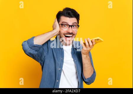 Emotional caucasian guy with glasses, hold smartphone in hand, screaming during phone call or recording audio message, stand on isolated orange background, holds his hand near head. Audio messaging Stock Photo