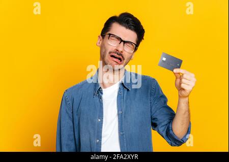 Crying upset caucasian unshaven stylish guy, holding a bank black card in his hand, bankrupt, debt, no money, upset, standing on isolated orange background, negative facial expression Stock Photo