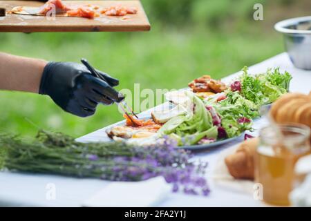 Male hands in gloves putting salmon from cutting board using iron twizzers. Side view of unrecognizable chef finishing dish, bread and fresh salad on plates, table decorated with lavender bouquet. Stock Photo