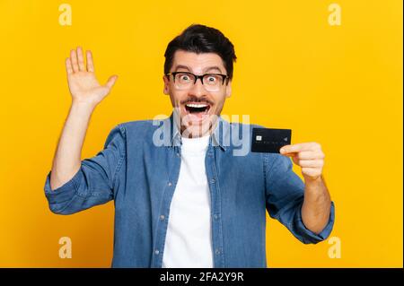 Young excited positive caucasian stylish man with glasses, holds credit card, rejoices in success, victory, profit, standing on isolated orange background happily raises his hand and smiling Stock Photo