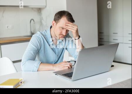 Sad unemployment young man sits in the kitchen at home and using laptop computer for searching opened vacant position, worried puzzled guy rests head on the hand Stock Photo
