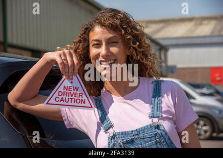 England, UK. 2021.  New driver, young attractive woman holding a new driver caution sign to display on her car after passing the driving test. Stock Photo