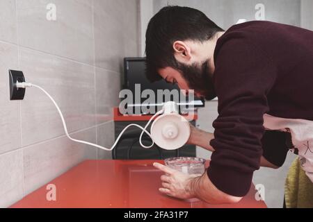 Young caucasian man in apron cooking in a kitchen. He is using a blender. Stock Photo