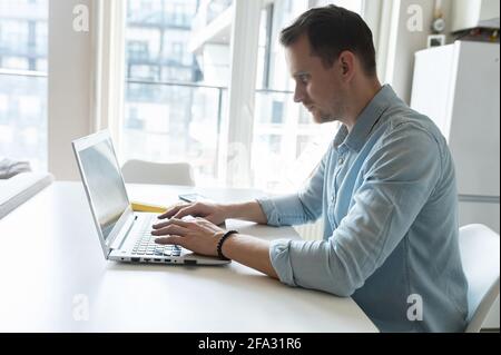Side view on concentrated young businessman typing on the keyboard, smart man wearing smart casual shirt answering emails, messaging online on the laptop working remptely from home Stock Photo