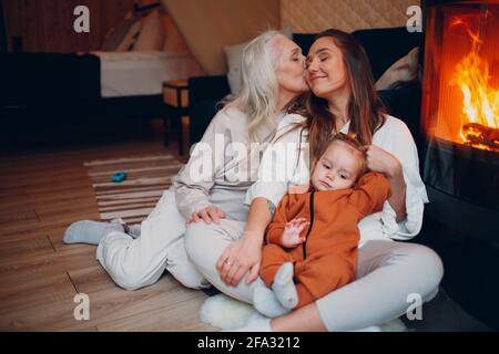 Grandmother, Mother and child kissing and playing on sofa near fireplace. Mom and baby. Parent with daughter and grandson little kid relaxing at home. Family having fun together. Mother's day Stock Photo