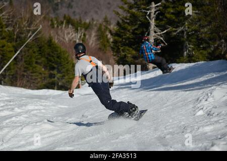 Two snowboarders seen from behind making a turn in Stowe Mountain resort in Vermont during Spring in mid-April warm sunny day. Stock Photo