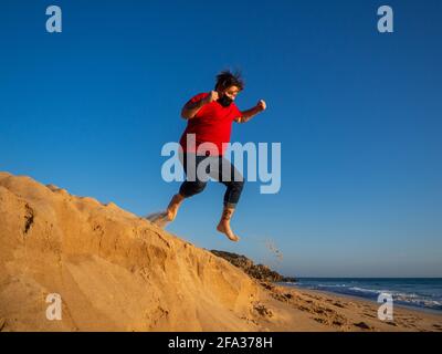 Spanish young man with a fabric mask jumping closer to the sandy coast against the clear blue sky Stock Photo