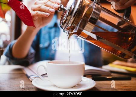 Woman's hands pouring tea into cup holding french press while sitting at table in cafe next to textbook and pen, close-up. Stock Photo