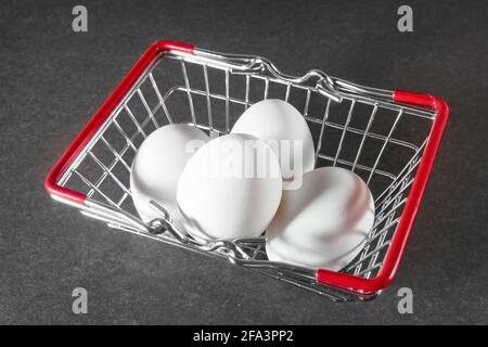Eggs, three white chicken eggs in small shopping trolley. Mini trolley with eggs on a dark gray background. Stock Photo