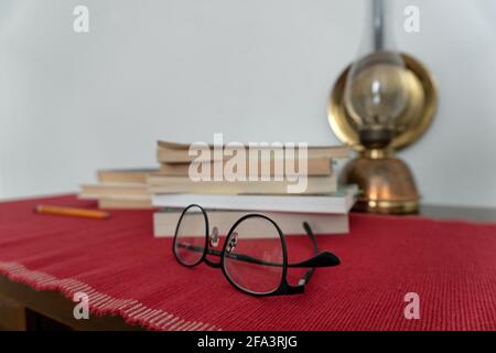 Eyeglasses in foreground and a book stack, a pencil on the left and an oil lamp in the background, lying on a red tablecloth. High quality photo Stock Photo