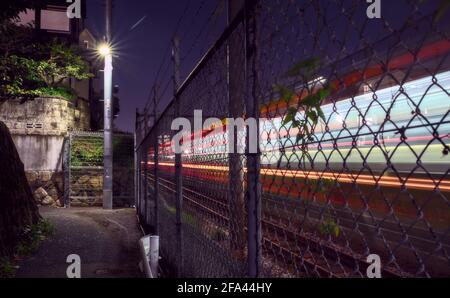 Long-exposure capture of light trails made by a train passing by an alleyway, separated from the tracks by a chain link fence Stock Photo
