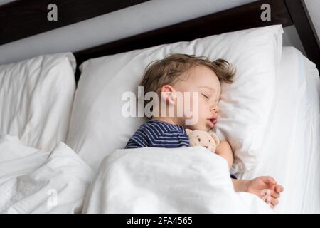 Authentic portrait sick cute caucasian little preschool baby boy in blue sleep with teddy bear on white bed. child resting at lunchtime. care Stock Photo