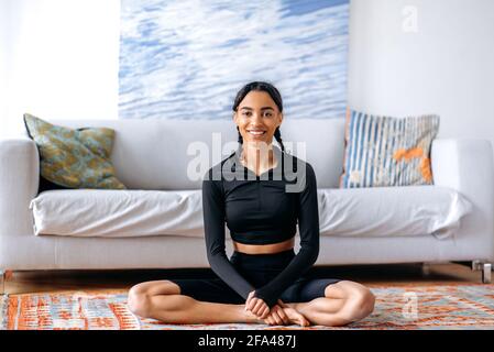 Relaxed young african american woman in black sportswear, sitting on the carpet at home in the lotus position, doing yoga, leading a healthy lifestyle, looking at the camera, smiling Stock Photo