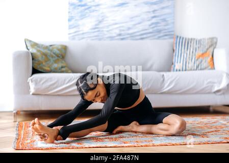 Flexible athletic young African American woman in sportswear, at home in the living room on the carpet, doing back stretching, doing yoga, leading a healthy lifestyle, closes eyes, smiling Stock Photo