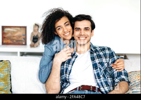 Portrait of happy mixed race young couple, african american girl tenderly hugs her beloved hispanic boyfriend, sitting on sofa, wearing casual stylish clothes, looking at camera, smiling Stock Photo