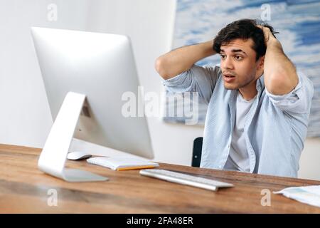 Surprised young hispanic businessman, freelancer or manager working at computer, sits at workplace, grabbed his head, had defeat, financial loss, business deal, looks anxiously at screen Stock Photo