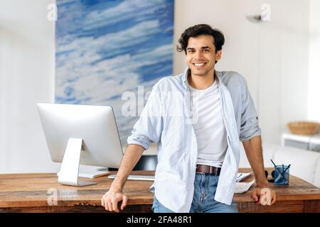 Portrait of confident successful young hispanic businessman, manager or freelancer, dressed in stylish clothes, standing near work desk, looking at camera, smiling friendly Stock Photo