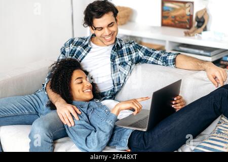 Young mixed married couple relaxing on sofa wearing casual clothes, african american girl lying on her boyfriend, using laptop, watching vacation photos or funny video, texting with friends, smiling Stock Photo