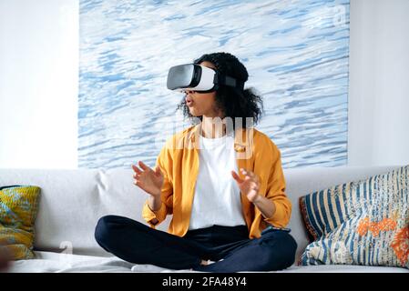 Excited African American girl sitting on a sofa at home in casual clothes, uses a virtual reality device, in a digital world, gesturing with her hands. Playing in the virtual world Stock Photo