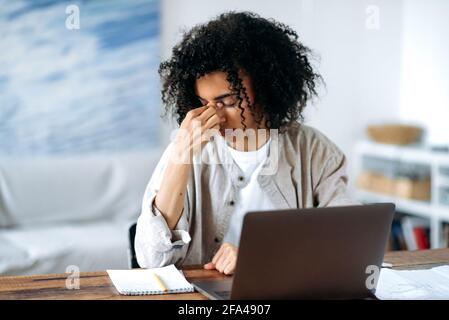 Tired frustrated overworked African American young woman in casual wear, manager, freelancer or student sitting in living room at laptop, experiencing stress, headache, rubs and close her eyes with Stock Photo