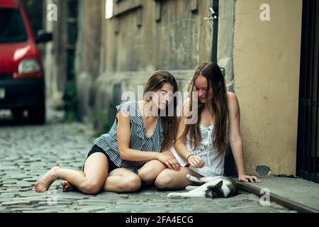 Two teenage girls sitting on the pavement with a cat. Stock Photo