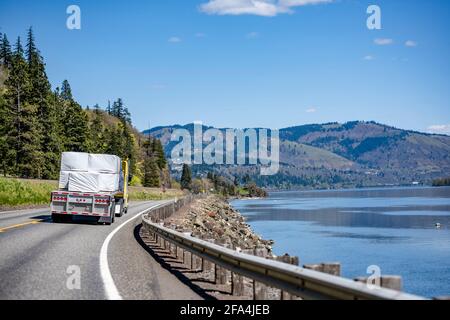 Bright yellow classic big rig semi truck transporting flat bed semi trailer loaded with covered lumber cargo turning on the winding highway road at na Stock Photo