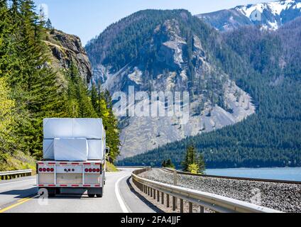 Bright yellow classic big rig semi truck transporting flat bed semi trailer loaded with covered lumber cargo turning on the winding highway road at na Stock Photo
