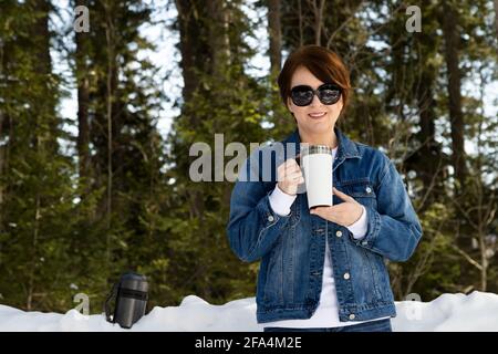 Mockup of a woman wearing sunglasses holding a white travel mug by a snowy woods. Empty travel coffee mug with handle template, model mockup Stock Photo