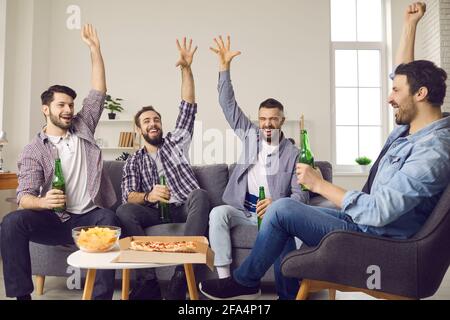 Smiling excited men group drink beer eat show hands up sit on home sofa Stock Photo