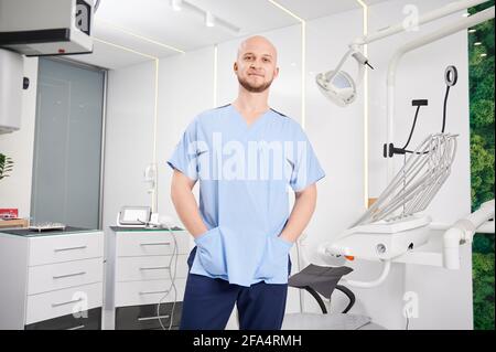 Portrait of handsome young man dentist looking at camera and smiling while keeping hands in pockets, standing in dental office with modern equipment. Concept dentistry, stomatology and dental workers. Stock Photo