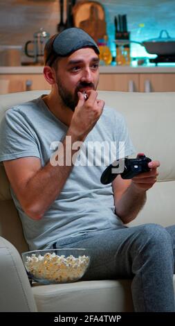 Upset pro gamer sitting on couch and playing soccer videogames for online competition using gaming joystick. Disappointed man looking at television eating popcorn late at night in kitchen Stock Photo