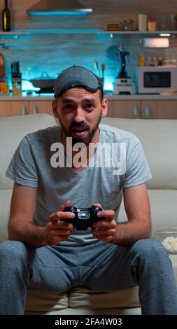 Upset pro gamer sitting in front of television losing soccer video games holding wireless controller. Frustrated man with sleep mask lying on sofa late at night in kitchen Stock Photo