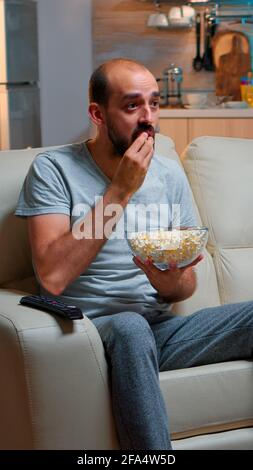 Sport football fan watching game late at night while enjoying popcorn, cheering for favourite football team. Championship entertainment match alone television, soccer fan looking at competition Stock Photo