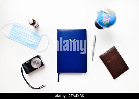 Top view travel accessories on white isolated background: blue notes book, globe stand, passport, compact camera, facial mask, sanitizer bottle. Stock Photo