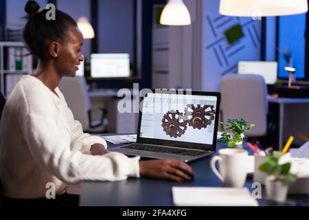 African engineer working late at night on a 3D model of industrial gears at laptop doing overtime in start up office. Freelancer studying prototype idea on pc showing cad software on device display. Stock Photo