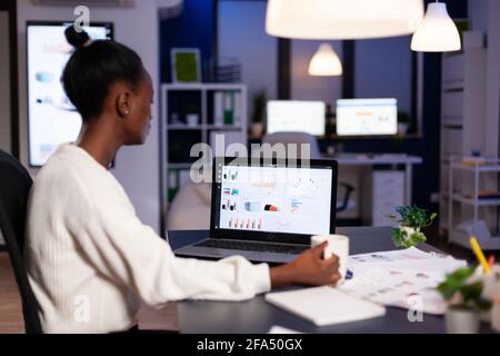 African businesswoman working with financial stats late at night in the office. Focused employee doing overtime for job respecting deadline of project writing, searching. Stock Photo