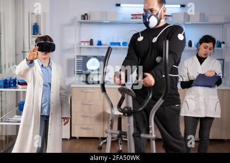 Scientist doctor in sport science laboratory wearing virtual reality goggles while athlete running, with electrodes attached on body monitoring physical endurancewhile ekg scan runs on computer screen. Stock Photo
