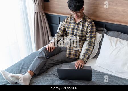 work from home, A Checkered shirt man who wearing eye glasses lays on the bed while using computer, whole body. Stock Photo