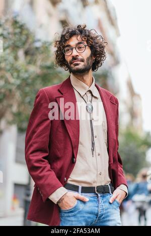 portrait of an attractive caucasian young man standing in the streets of Barcelona with his hands in his pockets. He has curly hair, beard and glasses. Dressing trendy casual clothes Stock Photo